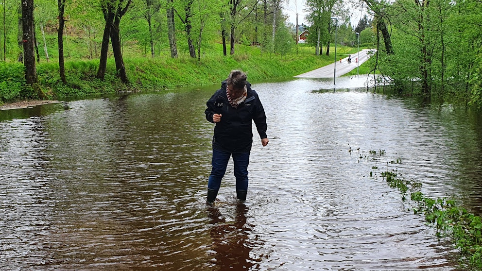 Reporter står i vatten vid översvämmad väg.