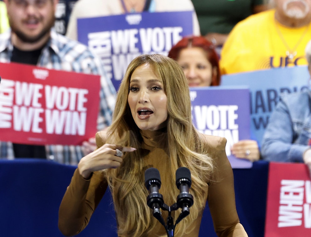 Skådespelerskan och sångerskan Jennifer Lopez klädd i brun polotröja står framför mikrofoner med Harrisväljare som håller upp skyltar i bakgrunden. 