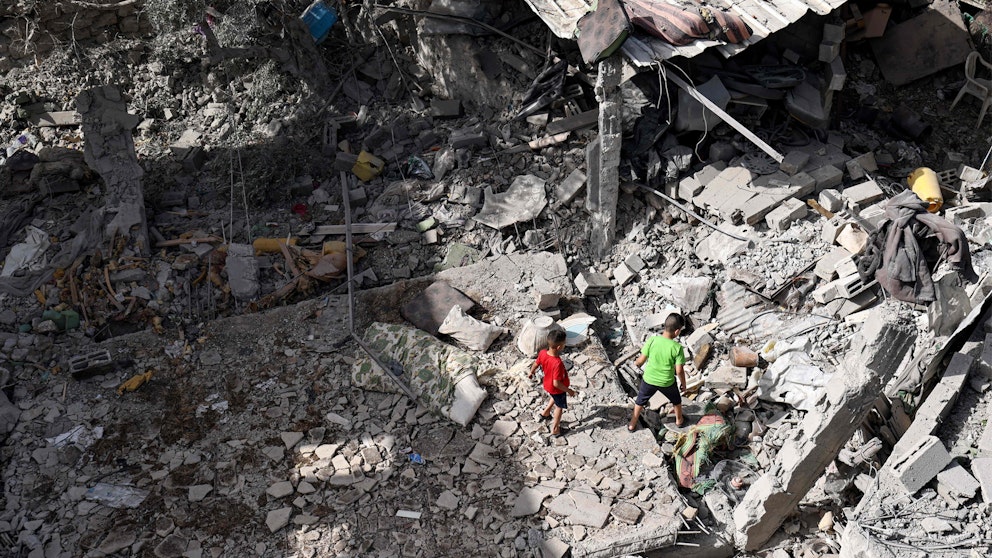 Två barn i en röd och en grön t-shirt syns bland resterna av en byggnad som jämnats med marken.