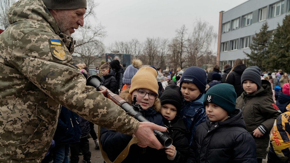 En stor samling barn kollar på ett vapen som hålls upp av en man i militärkläder. 