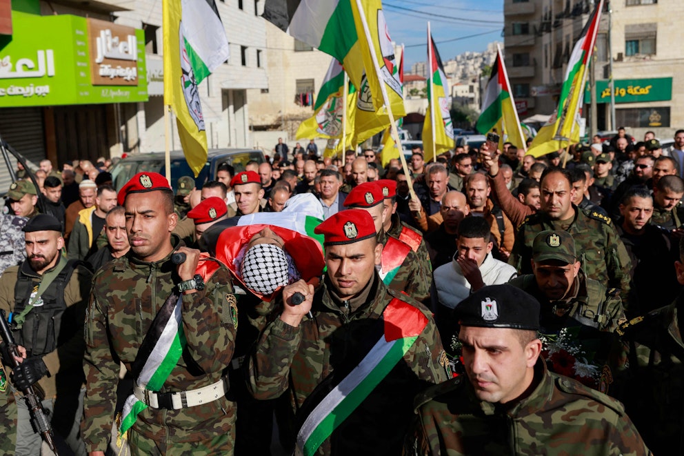 Män i röda baskrar och kamouflagemönstrade uniformer bär en död man med palestinasjal mellan vita hus på en gata full med folk. Ovanför syns den palestinska Fatahrörelsens flaggor i stort antal.