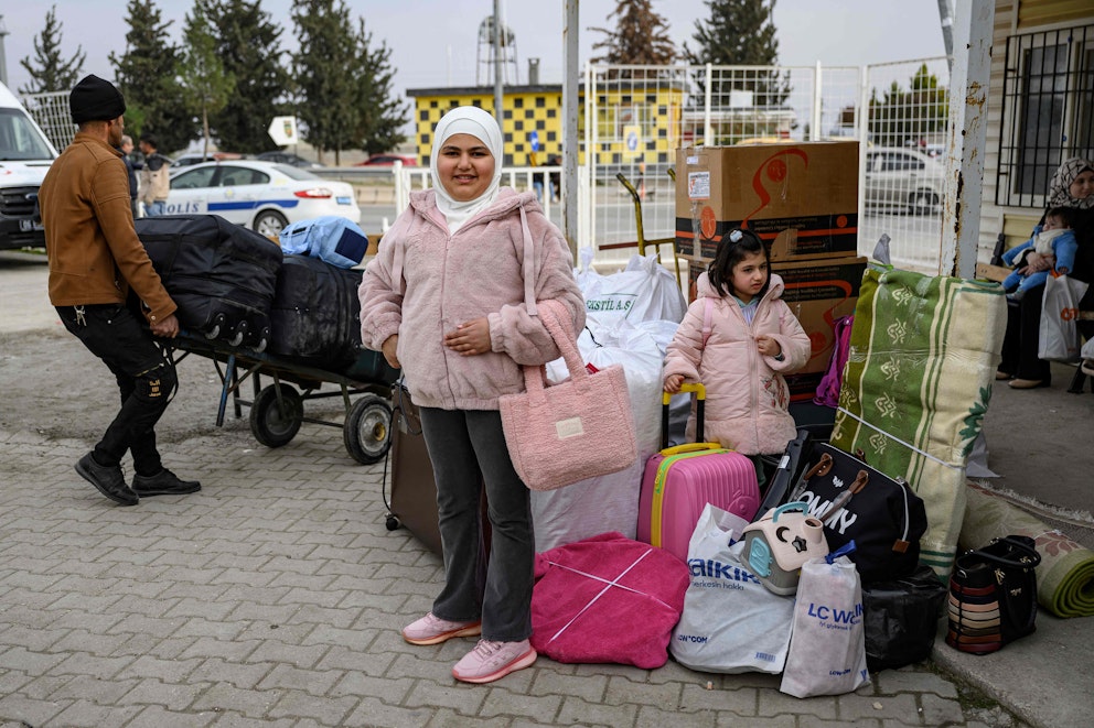 En person står framför en hög med bagage och kartonger på en gata, medan en annan person drar en kärra med mer bagage vid en gränsövergång.