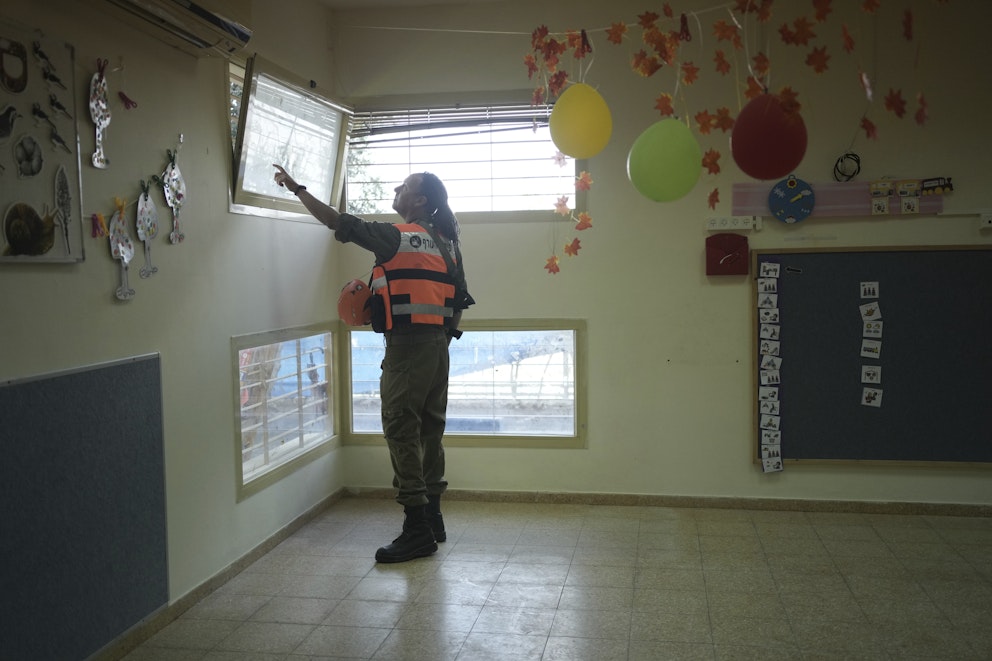 En uniformerad person står inuti ett rum med en säkerhetsväst och pekar uppåt mot ett fönster. Rummet är dekorerat med färgglada ballonger och pappersblad som hänger från taket. Väggarna är prydda med barnkonstverk och en tavla med bilder hänger på en av väggarna. Det finns flera fönster som släpper in naturligt ljus i rummet.