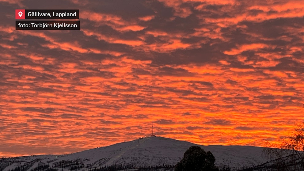 En spektakulär solnedgång färgar himlen djupt röd och orange över en snötäckt bergstopp i Gällivare, Lappland. De mörka konturerna av fjället framhävs mot de lysande molnen, medan sändarmasterna på toppen syns tydligt i silhuett.