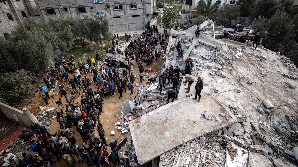 Många människor står vid en sönderbombad byggnad. Flera letar efter överlevare i rasmassan.