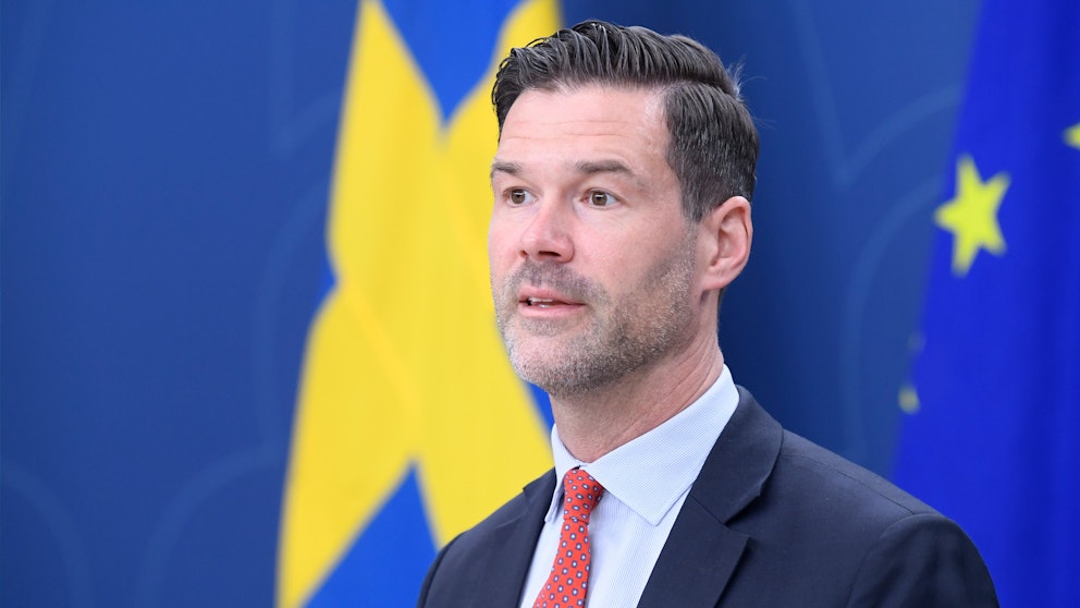 En man i kostym framför en svensk flagga och blå bakgrund.